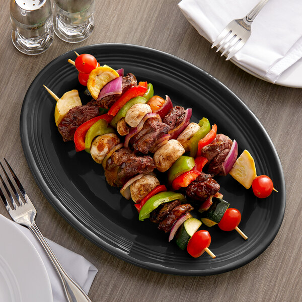 A Tuxton black oval china platter with meat and vegetables on skewers.