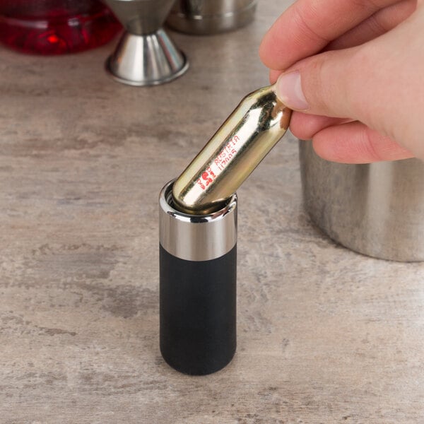 A hand holding a gold iSi CO2 soda charger over a black and silver metal cylinder.