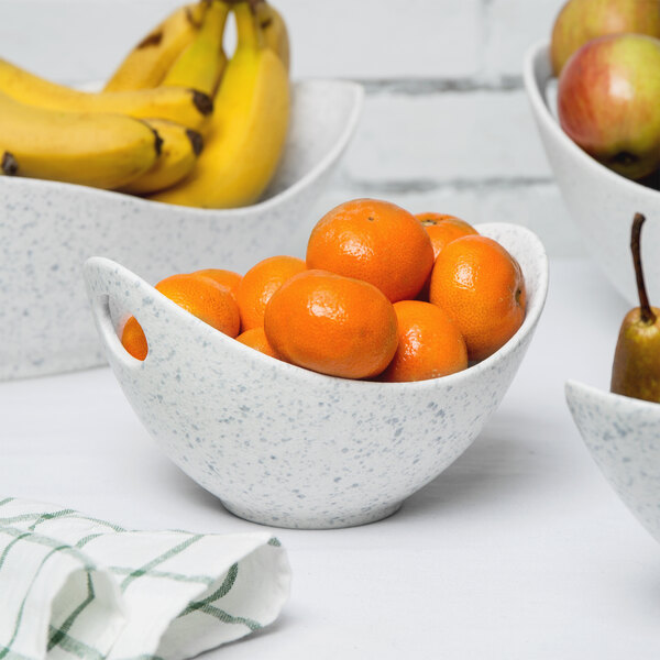 A white bowl with blue speckles and cut-out handles filled with oranges and bananas.