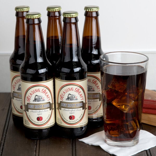 A group of Reading Soda Works Diet Root Beer bottles next to a glass of brown soda with ice cubes.