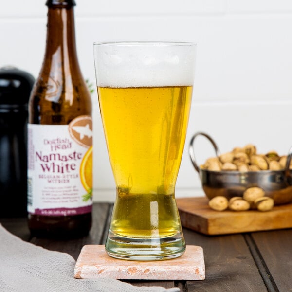 A Libbey pilsner glass of yellow beer sits on a wooden coaster next to a bottle of peanuts.