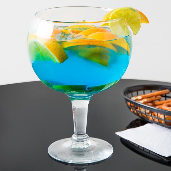 A Libbey Super Schooner glass with blue liquid and orange slices on a table with a bowl of pretzels.
