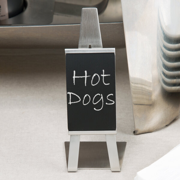 A stainless steel Tablecraft vertical card holder on a counter with a small black sign that says hot dogs.