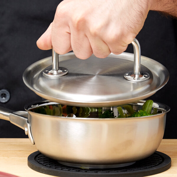 A hand holding a Vollrath stainless steel pot with a lid over a stove.