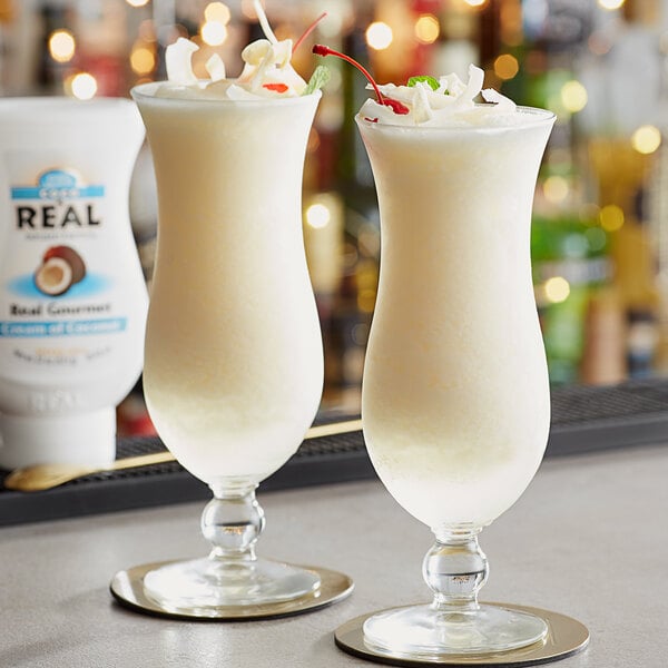 A bottle of Coco Real Cream of Coconut next to two glasses of tropical drinks topped with whipped cream and cherries.
