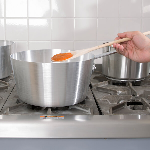 A person stirring Vollrath Arkadia sauce in a pot on a stove with a wooden spoon.
