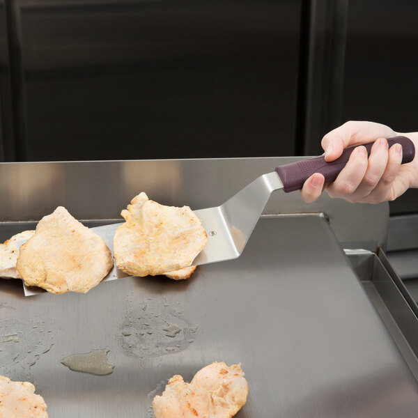 A Mercer Culinary Hell's Handle square edge turner with a purple handle being used to cut meat on a grill.