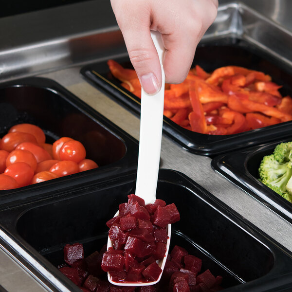 A hand holding a white Cambro perforated salad bar spoon of food over a container of vegetables.