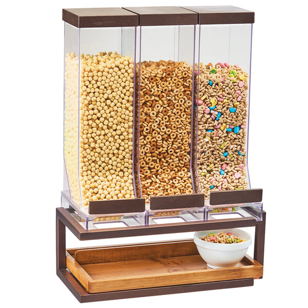 A Cal-Mil bronze metal and pine cereal dispenser with three containers holding a variety of cereals.
