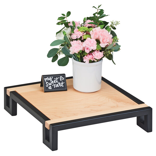A Cal-Mil metal framed maple display riser on a table with a white pot of pink flowers.