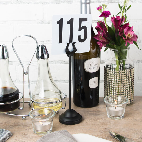 A black American Metalcraft harp table card holder with a number and flowers in a glass vase on a table.