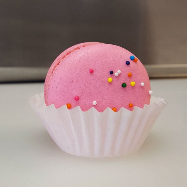A pink macaron with sprinkles on top sits in a white fluted mini baking cup.