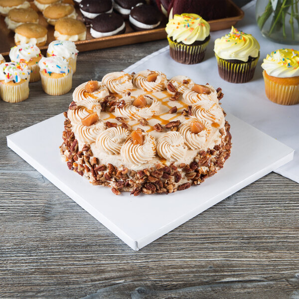 A white square cake drum with a cake with frosting and pecans on a table with cupcakes.
