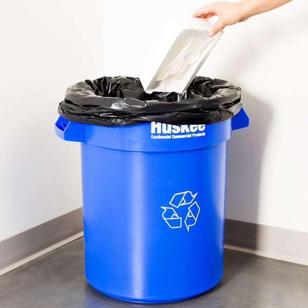 A person putting a black plastic bag into a blue Continental Huskee recycling bin.