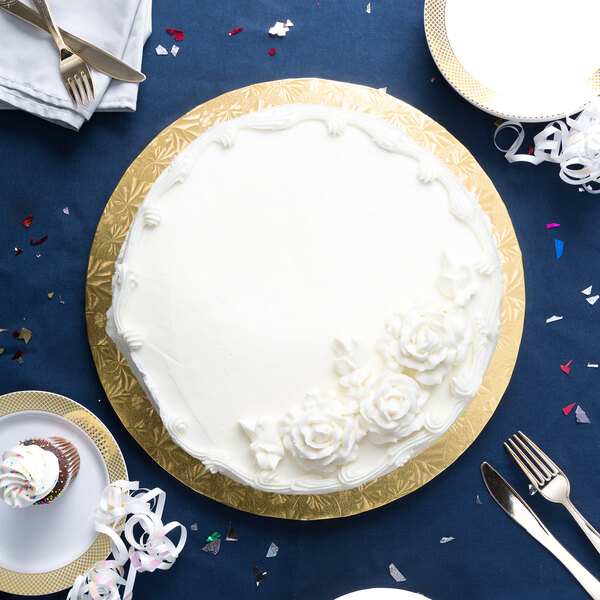 A white cake on a gold Enjay round cake drum with white frosting on a blue table with silverware