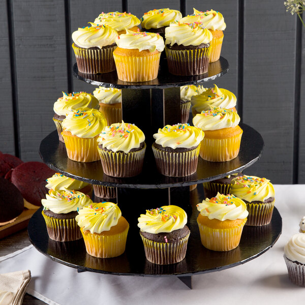 A Enjay black disposable cupcake stand holding three cupcakes with yellow frosting and sprinkles.