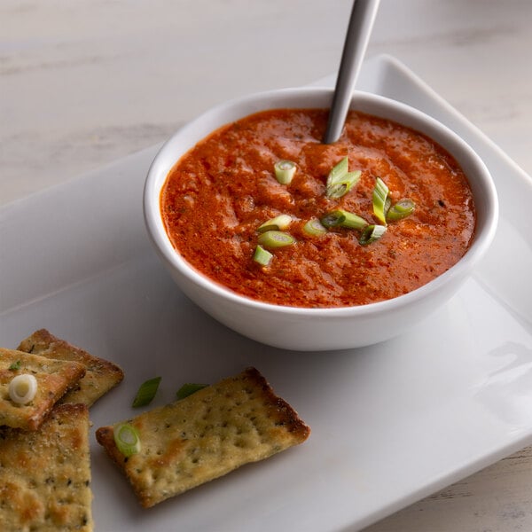 A bowl of tomato soup with a spoon and crackers on a plate.
