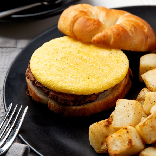 A breakfast sandwich with a croissant on a black plate with potatoes.