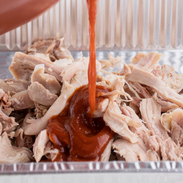 A person pouring sauce over Southern Hens Fully Cooked Pulled Chicken in a tin container.