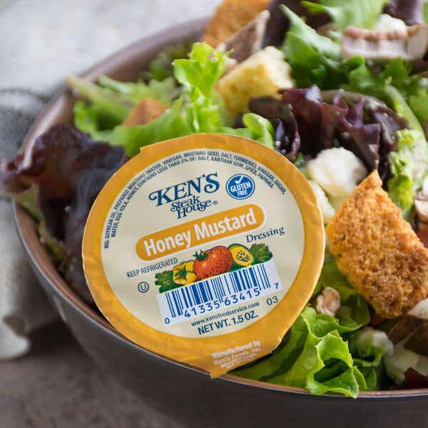 A bowl of salad with a round container of Ken's Foods Honey Mustard dressing.
