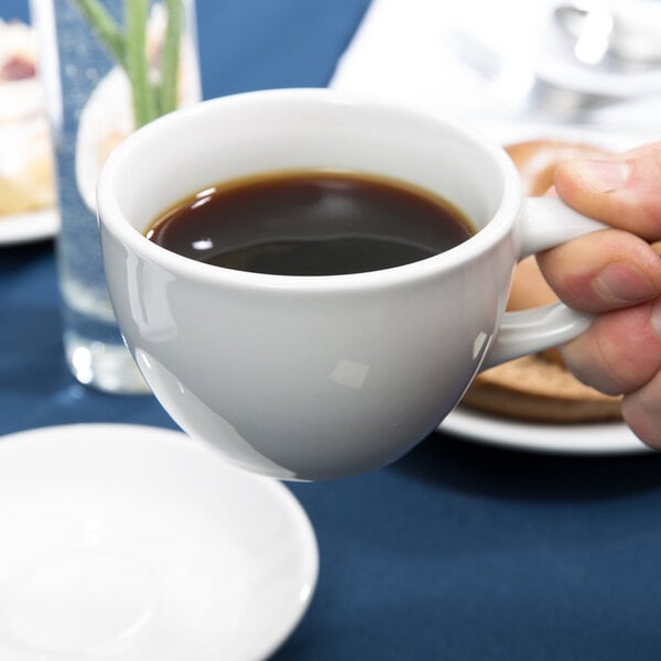 A person's hand holding a Tuxton Alaska white china cup filled with coffee over a white plate.