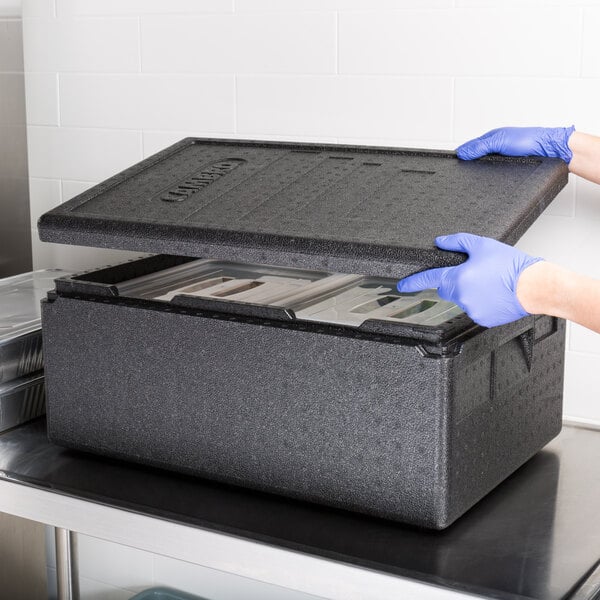 A person in blue gloves holding a black Cambro Cam GoBox with the lid open.