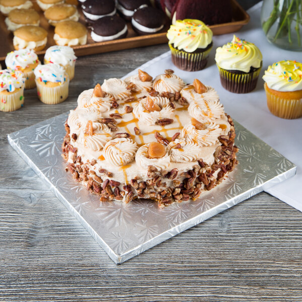 A cake with frosting and pecans on a silver square cake drum sitting on a table with cupcakes.