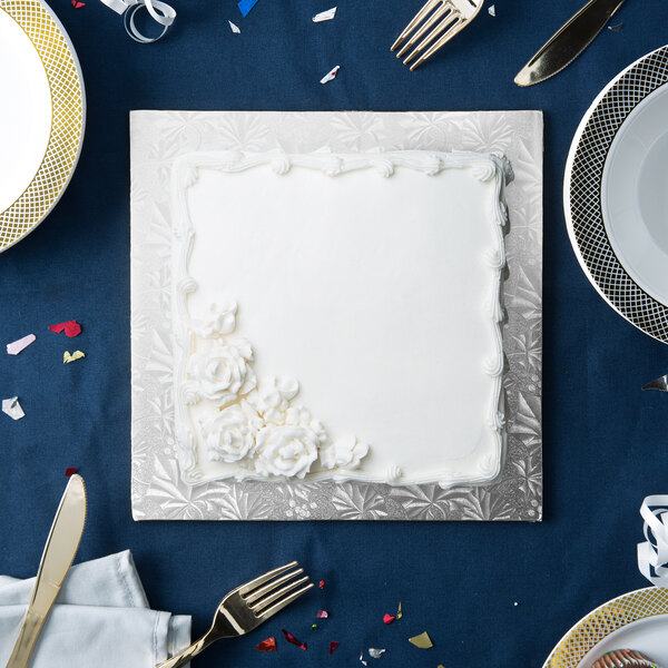 A white cake on a silver square cake drum on a blue table with silverware.