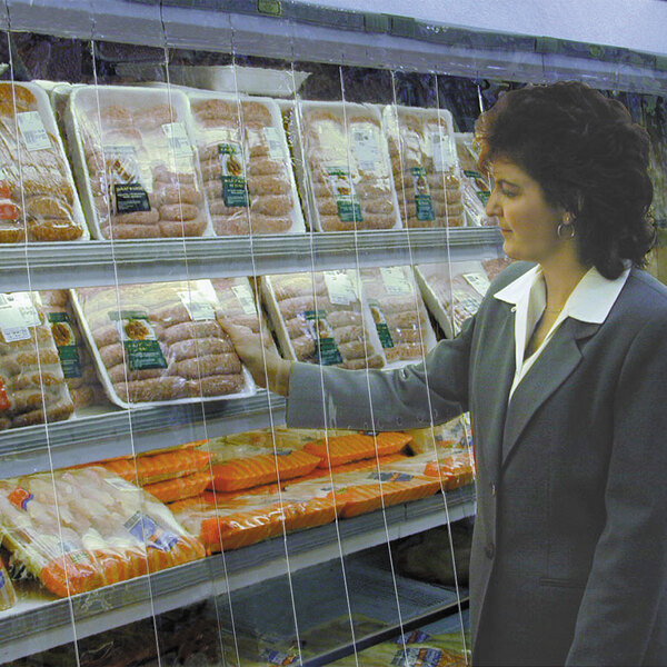 A woman looking at a display cooler with meats through a plastic strip curtain.