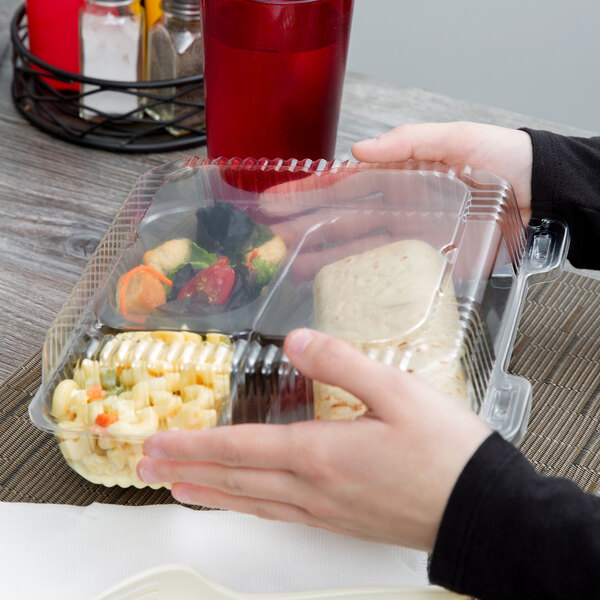 A hand holding a Durable Packaging clear hinged lid plastic container with three compartments of food.