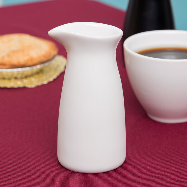 An Arcoroc white creamer on a table next to a cup of coffee.