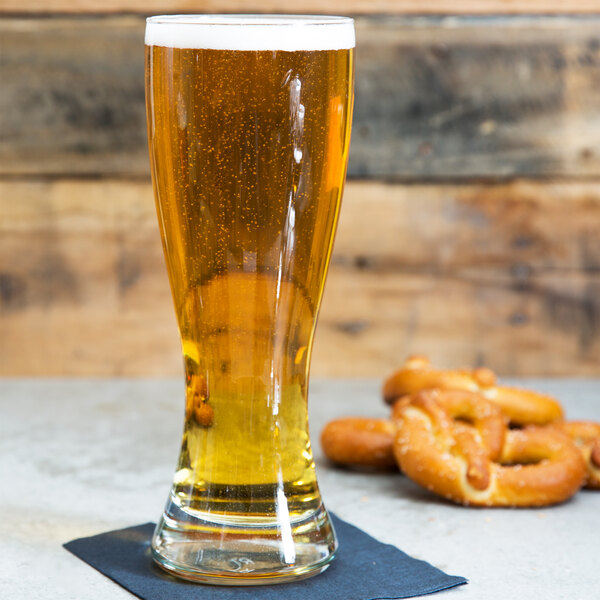 A close up of an Anchor Hocking Pilsner glass of beer next to a pretzel.