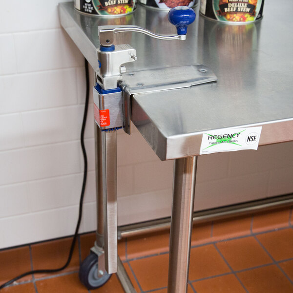 An Edlund Standard Duty manual can opener on a metal table with cans of food.