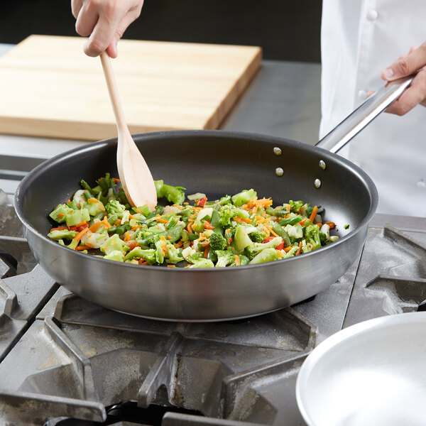 A person cooking broccoli and carrots in a Vollrath Centurion stainless steel non-stick fry pan.