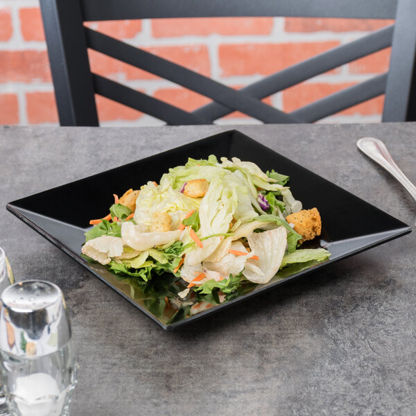 A black Siciliano square plate with a salad on it on a table.