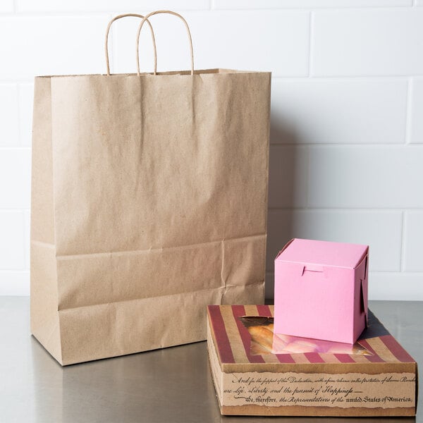 A brown paper bag with handles on a table next to a pink box.