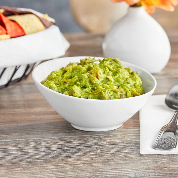 A slanted white porcelain bowl filled with guacamole on a table with a bowl of chips.