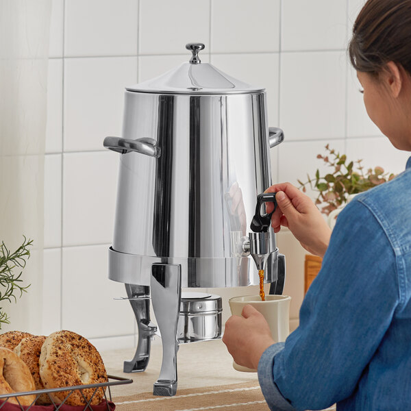 A woman pouring coffee into a Choice Deluxe stainless steel coffee chafer urn on a counter in a professional kitchen.