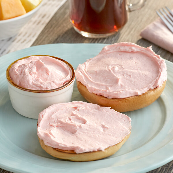 A plate with a bagel topped with strawberry cream cheese next to a bowl of fruit.