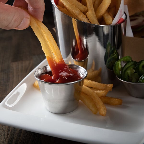A person dipping a french fry into a stainless steel sauce cup filled with ketchup.