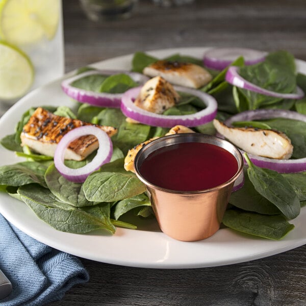 A plate of spinach salad with chicken and onions and a Choice copper-plated stainless steel sauce cup on a table.