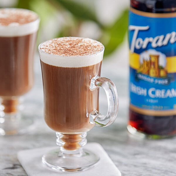 A glass mug of coffee with brown liquid and foam next to a Torani Sugar-Free Irish Cream Flavoring Syrup bottle.