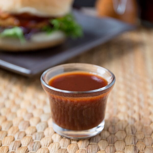 A small glass cup of Ken's Cannonball BBQ Sauce on a table next to a burger.
