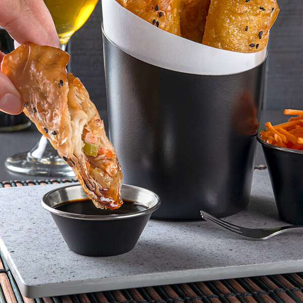 A hand holding a piece of food over a black sauce cup on a table.