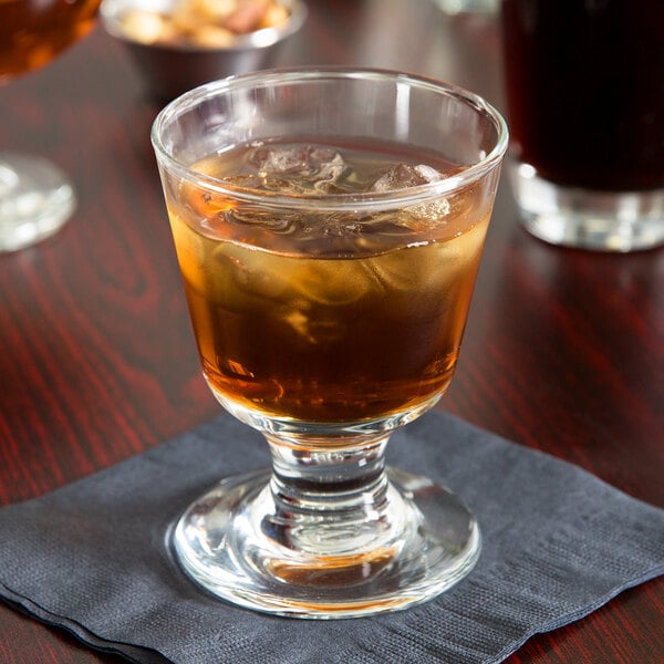 A Libbey footed rocks glass filled with brown liquid and ice on a napkin.