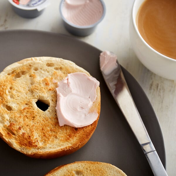 A plate with a bagel and a Philadelphia strawberry cream cheese cup next to a cup of coffee.