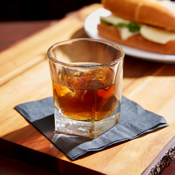 A Libbey Inverness rocks glass filled with brown liquid and ice sits on a napkin next to a sandwich.