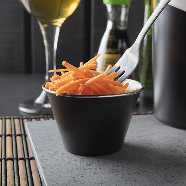 A close-up of a matte black stainless steel sauce cup with a fork and a carrot on it.