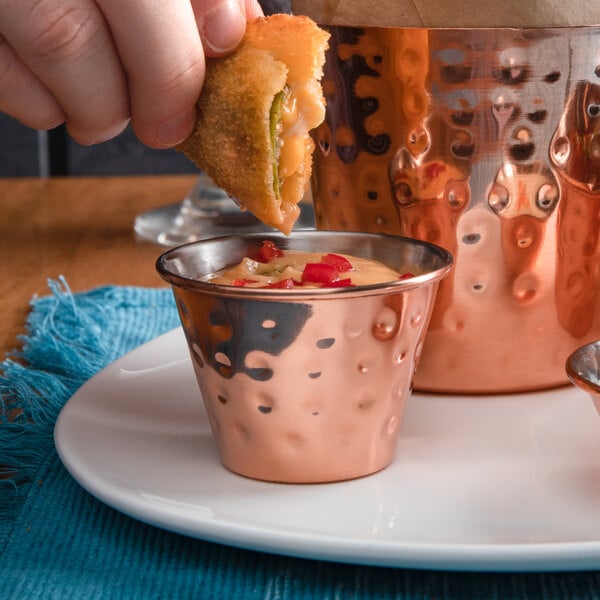 A person's hand dipping a piece of food into sauce in a copper cup.