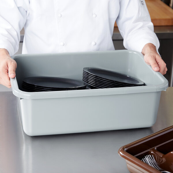 A chef in a white coat holding a light gray Cambro bus tub filled with black plates.